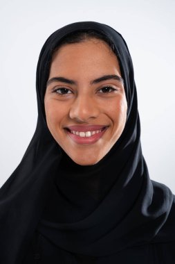Geleneksel siyah başörtüsü takan neşeli, genç bir Arap kadının stüdyo portresi temiz beyaz zemine karşı güven ve kültürel gurur sergiliyor.