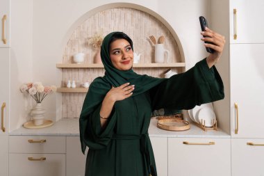 Gülümseyen emirati kadını şık bir mutfakta selfie çekiyor. Kurulumda aile ve ev yaşamına odaklı zarif bir dekor yer alıyor, kültürel ve kişisel ifadeleri vurguluyor