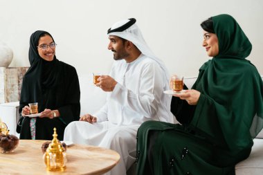 Geleneksel abayalar ve kanduralar giymiş Emirati dostları, modern bir kanepede dinlenirken aynı zamanda çayın tadını çıkarıyor ve neşeli sohbetler ediyor, bağlarını kutluyorlar.