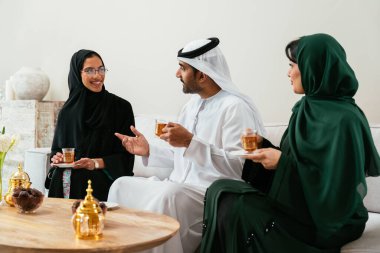 Geleneksel abayalar ve kanduralar giymiş Emirati dostları, modern bir oturma odasında canlı sohbetler ederken çay ve tarihlerin tadını çıkarıyorlar.