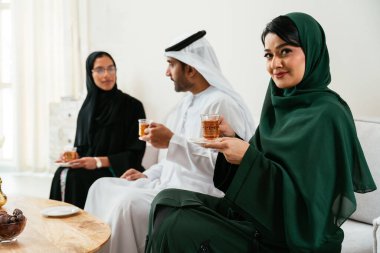 Geleneksel abayalar ve kanduralar giymiş Emirati 'li kadınlar ve erkekler, modern bir oturma odasında bir kanepede oturmuş çay ve sohbetin tadını çıkarıyor, dostluklarını ve kültürel miraslarını kutluyorlar.