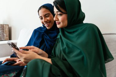 Geleneksel abayalar giyen iki emirati kadını, modern bir oturma odasında rahat bir koltukta oturuyorlar, akıllı telefona bakarken bir anlık neşe ve kahkaha paylaşıyorlar.