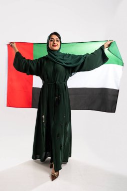 Geleneksel yeşil abaya giyen Emirati kadını ve tesettür gururla bir stüdyo ortamında ulusal gurur ve kültürel kimliği somutlaştıran uae bayrağını tutuyor.