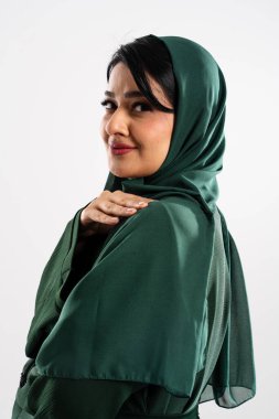 Geleneksel yeşil başörtüsü takan gülümseyen müslüman bir kadının stüdyo portresi omzunun üstünden beyaz bir arka plana bakarken nazikçe omzuna dokunuyor.