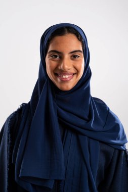 Mavi tesettürlü neşeli genç bir Arap kadının stüdyo portresi, temiz beyaz bir arka planda güzellik ve kültürel kimliğini sergiliyor.