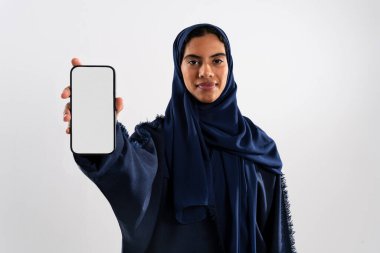 Geleneksel mavi abaya ve tesettür giyen bir Arap kadının stüdyo portresi, önünde boş ekranla akıllı bir telefon tutuyor, bir uygulama veya web sitesini tanıtıyor