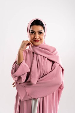 Pembe bir abaya ve tesettüre zarif bir şekilde poz veren özgüvenli ve şık bir müslüman kadının stüdyo portresi, minimalist beyaz bir arka plana karşı zarafet ve kültürel gurur yayıyor.