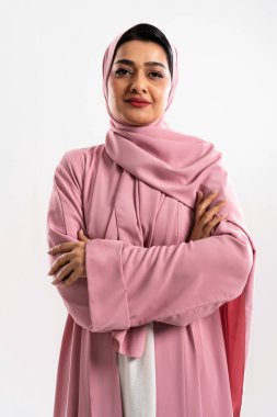Pembe bir abaya ve tesettür giyen, beyaz bir arka planda çapraz kollarla duran, kendinden emin bir ortadoğulu iş kadınının portresi, profesyonellik ve kültürel gurur yayıyor.