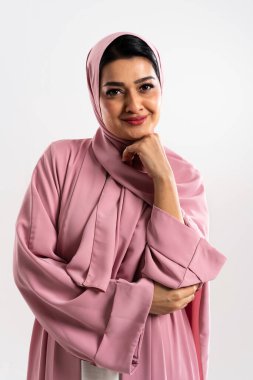 Pembe bir abaya ve tesettür giyen kendine güvenen ve şık bir müslüman kadının stüdyo portresi, eli nazikçe çenesine dokunuyor, zarafet ve zarafet saçıyor minimalist beyaz bir arka plana karşı.