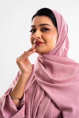Kendine güvenen, zarif, pembe tesettürlü bir Müslüman kadının stüdyo portresi. Elleriyle nazikçe yüzüne dokunuyor. Geleneksel ortadoğu moda ve kültürel güzelliği sergiliyor.