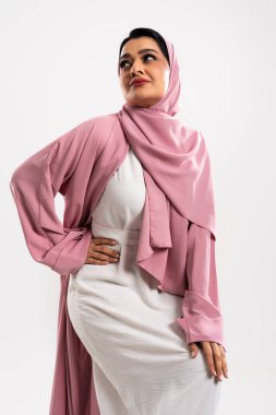 Pembe tesettür ve abaya giyen Arap kadın, modern İslami moda ve zarafeti sergileyen beyaz bir stüdyo arkaplanına karşı güvenle poz veriyor.