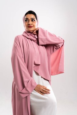 Pembe bir abaya ve tesettüre bürünmüş, özgüvenli ve modaya uygun bir ortadoğu kadınının stüdyo portresi, zarafet ve duruş ile modern İslami giysiler sergiliyor.
