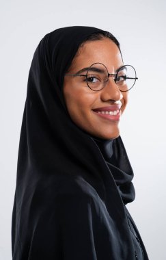 Gözlük takan gülümseyen bir Arap kadının stüdyo portresi ve geleneksel siyah bir tesettür, tarafsız bir arkaplan karşısında özgüvenini ve kültürel kimliğini gösteriyor.