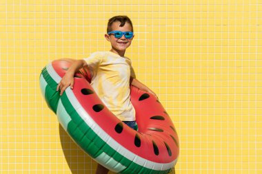 Mutlu ve havalı Çinli genç yaz mevsiminde sahilde eğleniyor. Asyalı bir çocuk sarı arka plan duvarında güneş gözlüğü ve mayo portresi takıyor.