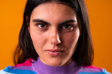 Uzun siyah saçlı ve çilli genç bir kadının stüdyo portresi. Renkli bir süveter giyiyor. Turuncu bir arka plana doğrudan ve yoğun bir şekilde bakıyor.