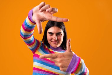 Renkli çizgili bir süveter giyen genç bir kadının stüdyo portresi parmaklarıyla canlı turuncu bir zemin oluşturarak yaratıcılık ve bireyselliği ifade ediyor.