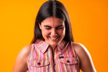 Uzun kahverengi saçlı neşeli genç bir kadın canlı turuncu bir arka plana karşı gözleri kapalı gülüyor, renkli çizgili kolsuz bir gömlek giyiyor ve neşe saçıyor.