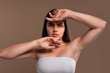 Çilli güzel bir genç kadının stüdyo portresi yüzünü çerçeveleyen elleriyle poz veren, doğal güzelliğini ve benzersiz özelliklerini vurgulayan beyaz bir bando bluzu giyerken.