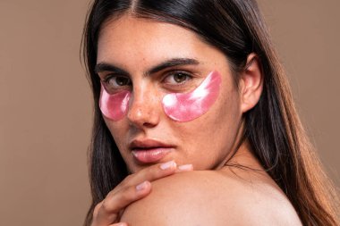 Uzun kahverengi saçlı ve çilli güzel bir kadının stüdyo portresi. Cilt bakımının bir parçası olarak pembe göz altı bantları uyguluyor. Güzelliği ve kendine bakımı teşvik ediyor.