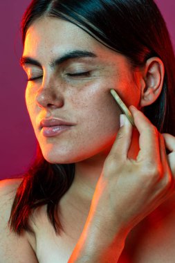 Gözleri kapalı genç bir kadın yüzüne cilt bakımı uyguluyor, doğal güzelliği ve kendine bakımı vurguluyor. Stüdyo aydınlatması onun eşsiz özelliklerini ve dingin ifadesini vurguluyor