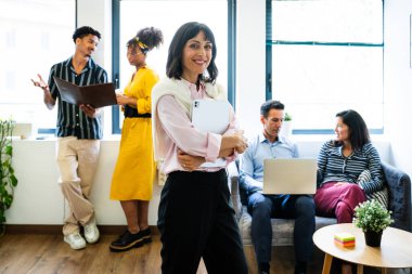 Kendine güveni tam bir iş kadını, çeşitli takımları tarafından çevrelenmiş, parlak, modern bir ofis girişimi ortamında işbirliği yapan ve çalışan, yenilik ve takım çalışmasını teşvik eden belgeleri tutarken gülümsüyor.