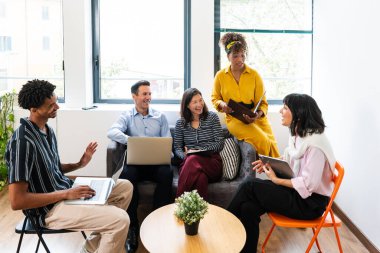 Beş gülümseyen iş adamı modern bir ofiste laptopları ve tabletleri kullanarak, fikirleri paylaşarak ve birlikte çalışarak verimli bir toplantı yapıyorlar.