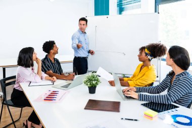 Çeşitli iş adamları bir toplantıya katılıyor, bir yöneticinin beyaz tahtada bir strateji sunuşunu dikkatle dinliyor, işbirliği ve yeniliği destekliyor.