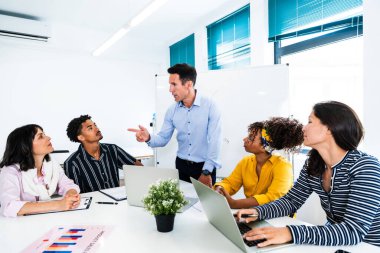 Profesyonellerden oluşan çeşitli takımlar parlak, modern bir ofiste masanın etrafında toplanarak, yöneticileri tarafından yönetilen ve bir beyaz tahtaya işaret eden bir beyin fırtınasına aktif olarak katılıyorlar.