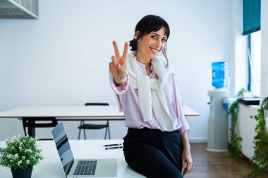 Neşeli bir ofiste, modern bir masada oturmuş gülümseyen iş kadını zafer işareti sergiliyor ve gelişen bir ortamda başarısını ve başarılarını kutluyor.