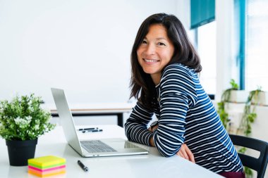 Bitkiler ve yapışkan notlarla dolu, parlak, modern bir ofiste çalışırken gülümseyen Asyalı işkadını, olumlu ve üretken bir çalışma ortamı sergiliyor.