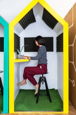 Genç iş kadını işine yoğunlaşırken renkli, ses geçirmez bir ofis kapsülünde dizüstü bilgisayar kullanıyor. Gizlilik ve modern bir çalışma alanına odaklanmak için tasarlanmış.