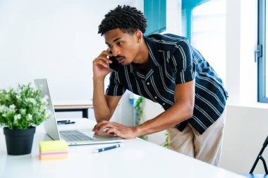 Günümüz iş dünyasında verimlilik ve bağlantıyı gösteren, modern ofiste dizüstü bilgisayarla çalışırken akıllı telefonlardan konuşan genç iş adamı.