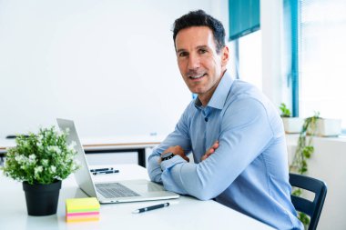 Masasında oturan gülümseyen bir iş adamının portresi kollarını kavuşturmuş, parlak, modern bir ofiste dizüstü bilgisayarla çalışıyor, güven ve profesyonellik yayıyor.