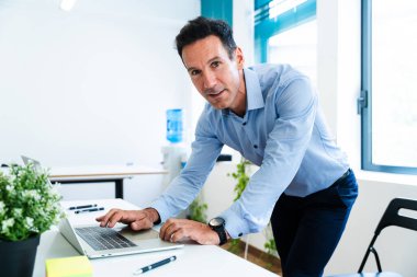 Kendine güveni tam bir iş adamı, parlak, modern bir ofiste dizüstü bilgisayarla çalışıyor, verimliliği ve profesyonel bir çevreye odaklanmayı öneriyor.