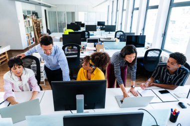 Parlak ve modern bir ofiste birlikte çalışan çeşitli profesyonellerden oluşan bir ekip, dizüstü bilgisayar ve bilgisayar kullanan bir proje üzerinde işbirliği yaparak, bir girişim ortamında takım çalışması ve üretkenliği gösteriyorlar.