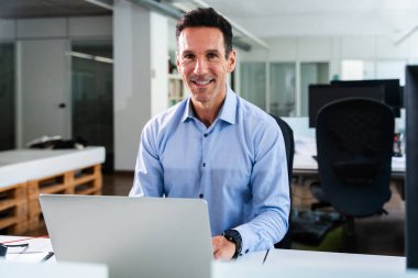 Parlak, modern bir ofiste dizüstü bilgisayarla çalışan gülümseyen işadamı, kariyerinin verimliliğinin ve başarısının keyfini çıkarırken, aynı zamanda pozitif bir çalışma ortamının tadını çıkarıyor.