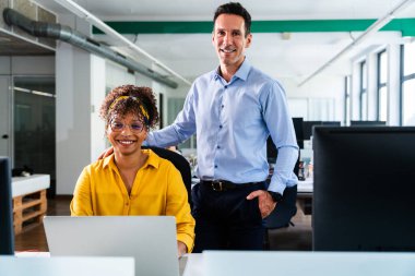 İki gülümseyen iş arkadaşı, dizüstü bilgisayar kullanan bir kadın ve cebi dolu bir adam, modern ofis ortamında birlikte çalışıyorlar, bir proje üzerinde çalışıyorlar, takım çalışması ve üretkenlik gösteriyorlar.