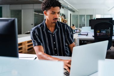 Genç işadamı, modern bir ofisteki dizüstü bilgisayarına yoğunlaşarak profesyonelliğini ve görevlerine olan bağlılığını gösteriyor.