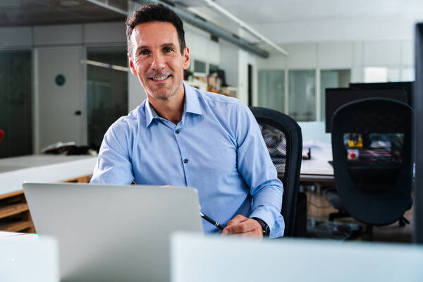 Зрелый бизнесмен сидит за своим столом, уверенно улыбаясь, работая над своим ноутбуком в яркой, современной офисной обстановке, держа ручку и источая профессионализм и успех