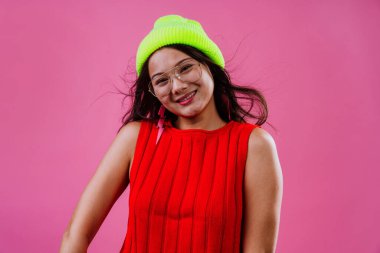 Canlı kırmızı bir bluz ve neon sarısı bir bere giyen, pembe bir arka plana neşeyle gülen, modern ve eğlenceli bir moda anlayışı sergileyen, etkileyici genç bir kadın.