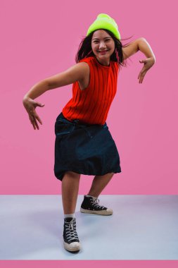 Canlı kırmızı bir bluz ve neon sarısı bir bere giyen, pembe bir arka plana neşeyle gülen, modern ve eğlenceli bir moda anlayışı sergileyen, etkileyici genç bir kadın.