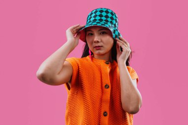 Kameraya gülümseyen genç bir kadın, turuncu örgü bir süveter ve canlı bir deniz mavisi ve siyah kareli bir kova şapka giyiyor, parlak pembe bir arka planda duruyor.