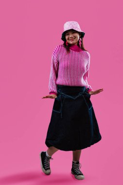 Pembe desenli kova şapkalı, çizgili kazaklı, neşeli pembe stüdyo arkaplanına gülümseyen ve kameraya bakan Asyalı kadın.