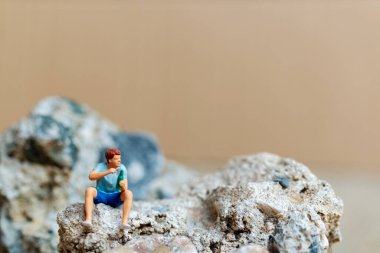 Minyatür insanlar, kayanın üzerinde otururken bira içip sigara içen genç bir adam.