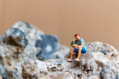 Minyatür insanlar, kayanın üzerinde otururken bira içip sigara içen genç bir adam.