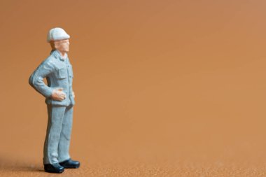 Minyatür inşaat işçisi. Mavi iş üniforması giymiş, kasklı bir adam. Elleri kalçasında, özgüvenle duruyor, hazır olma ve kararlılık hissini yansıtıyor.