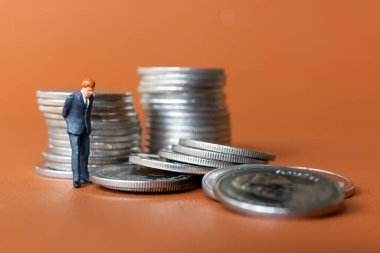 Minyatür insanlar. Takım elbiseli bir işadamı, turuncu arka planda birkaç gümüş para destesiyle ayakta duruyor..