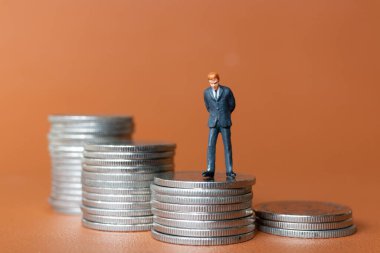 Minyatür insanlar. Takım elbiseli bir işadamı, turuncu arka planda birkaç gümüş para destesiyle ayakta duruyor..