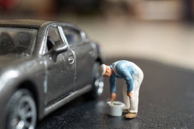 Minyatür insanlar, bir işçi, metalik gri bir arabanın arka planındaki bir şeyi inceler ya da tamir eder gibi eğilir.