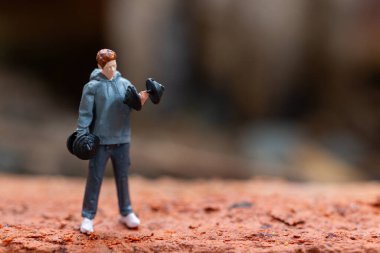 Minyatür insanlar, dambılları kaldıran bir adam desenli bir arka plana ayarlıdır. Gri kapüşonlu ve koyu renk pantolonlu figür, minyatür bir dünyada forma ve güce odaklanmayı temsil ediyor..